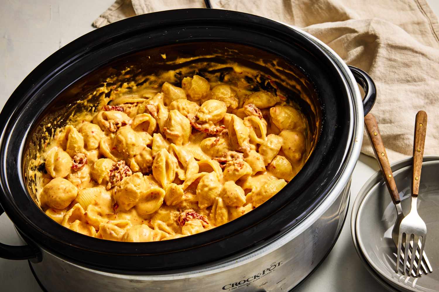 A slow cooker insert filled with creamy mac and cheese as a spoon lifts a stretchy, cheesy scoop on a kitchen counter in soft natural light