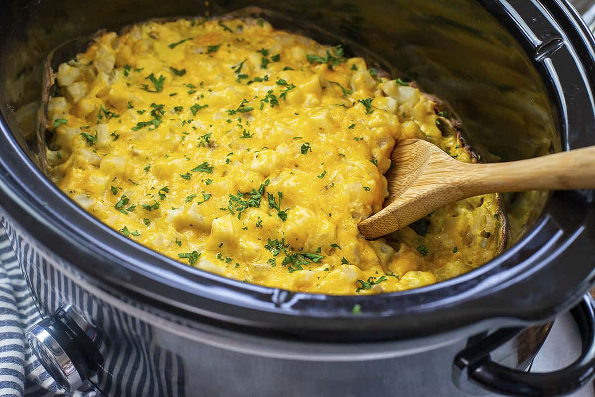 A slow cooker insert filled with frozen diced hash browns, chopped onion, garlic, and broth being stirred with a wooden spoon
