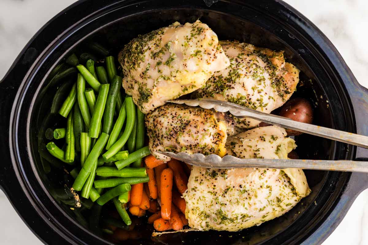 A slow cooker insert filled with raw chicken thighs, chopped carrots, celery, and onions with broth being poured in
