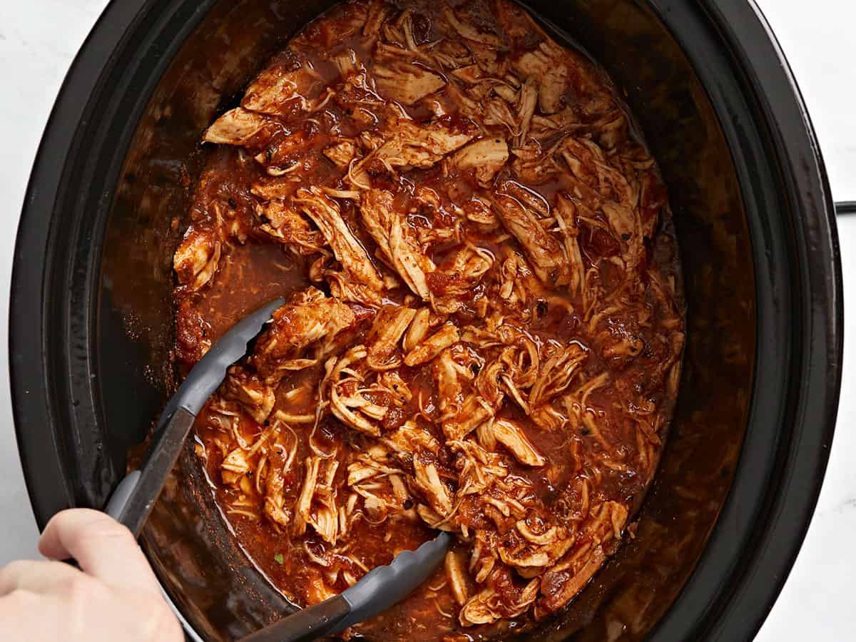 A slow cooker insert filled with tender shredded chicken in a red salsa sauce, with steam rising and a ladle resting on the edge on a bright kitchen counter