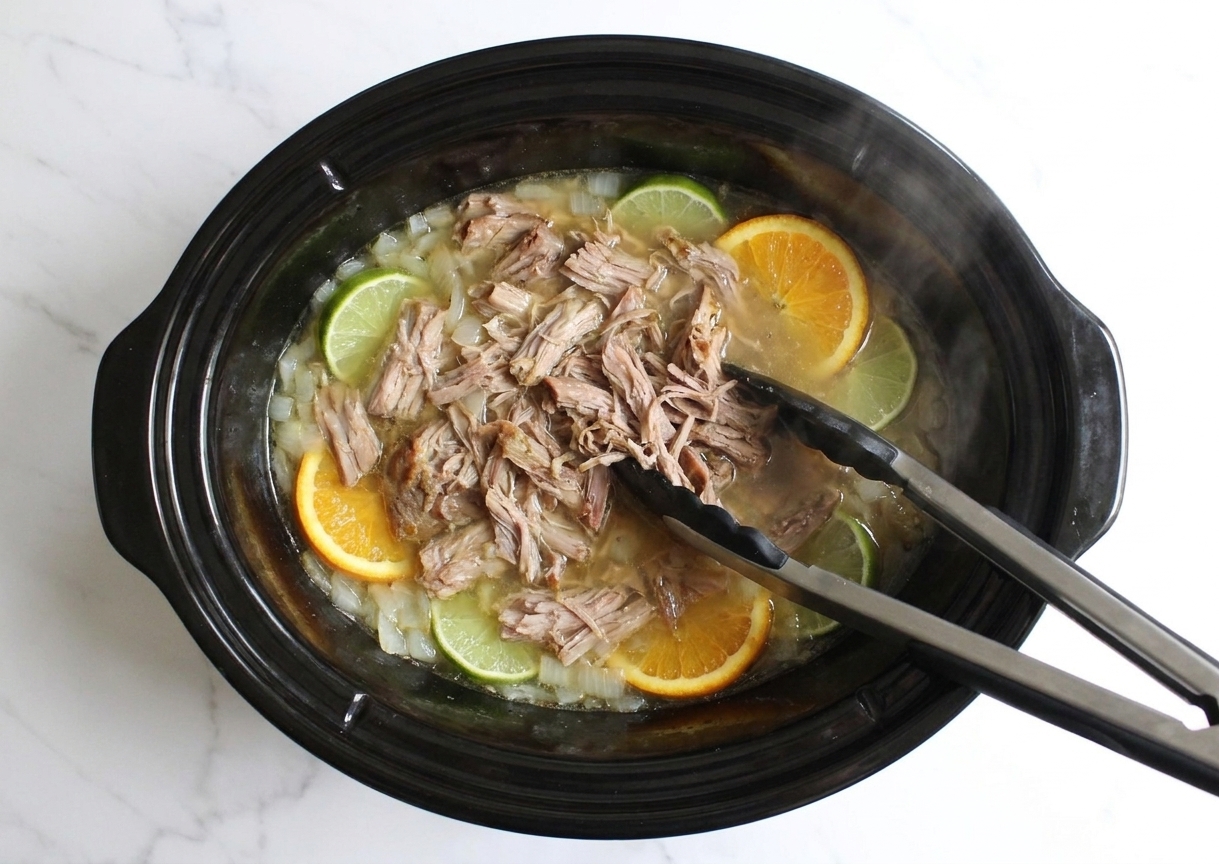 A slow cooker insert filled with tender shredded pork and onions in a light citrusy broth, with a pair of tongs pulling the meat apart