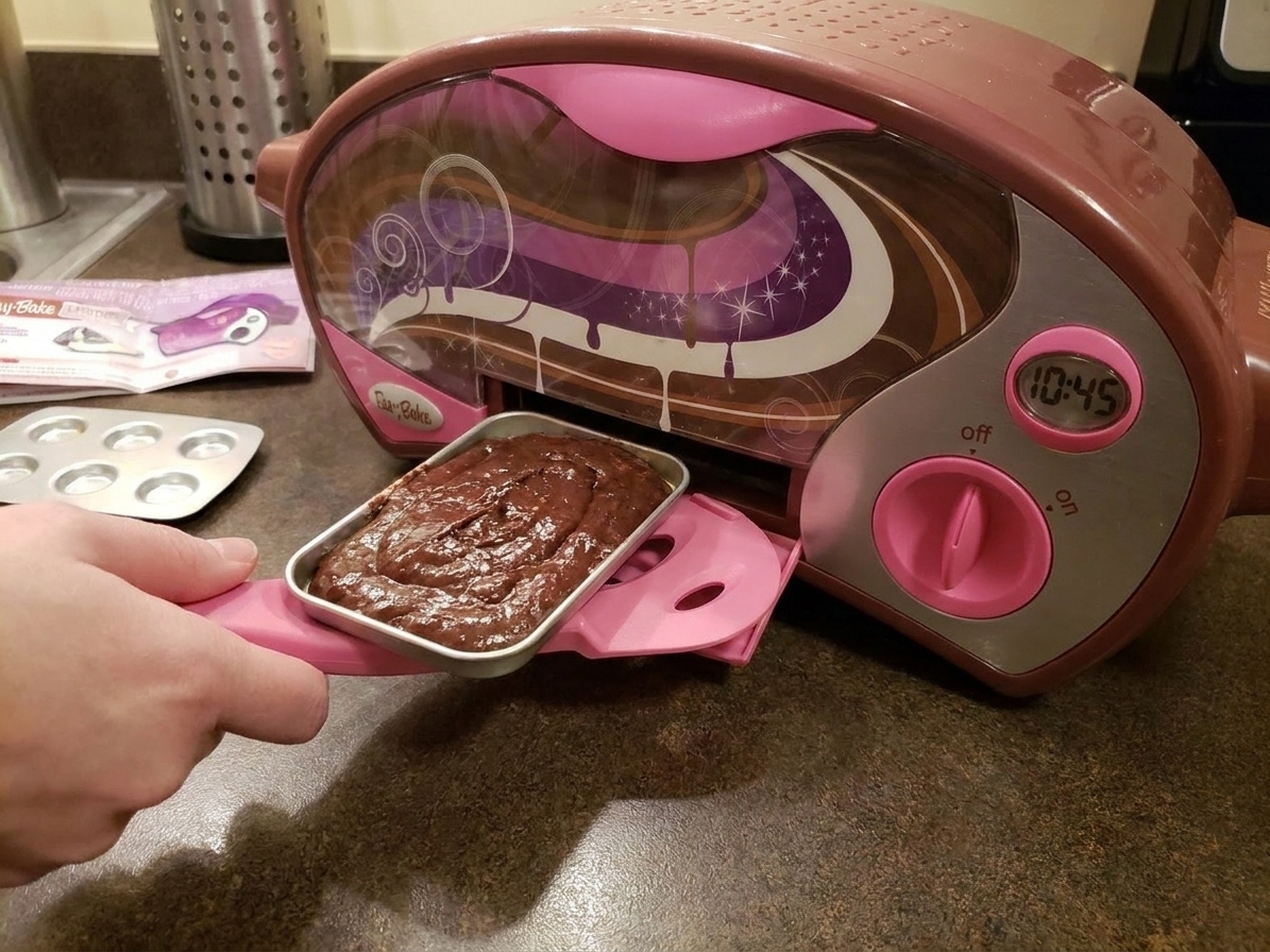 A small Easy-Bake style oven on a kitchen counter with the brownie pan being slid into the baking slot