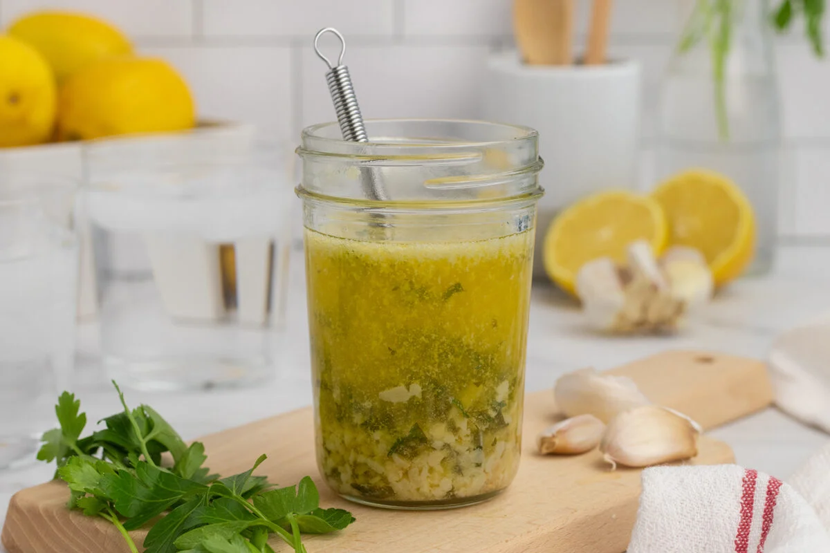 A small arrangement of butter, a whole lemon, minced garlic, chopped parsley, salt, pepper, and olive oil on a light kitchen counter