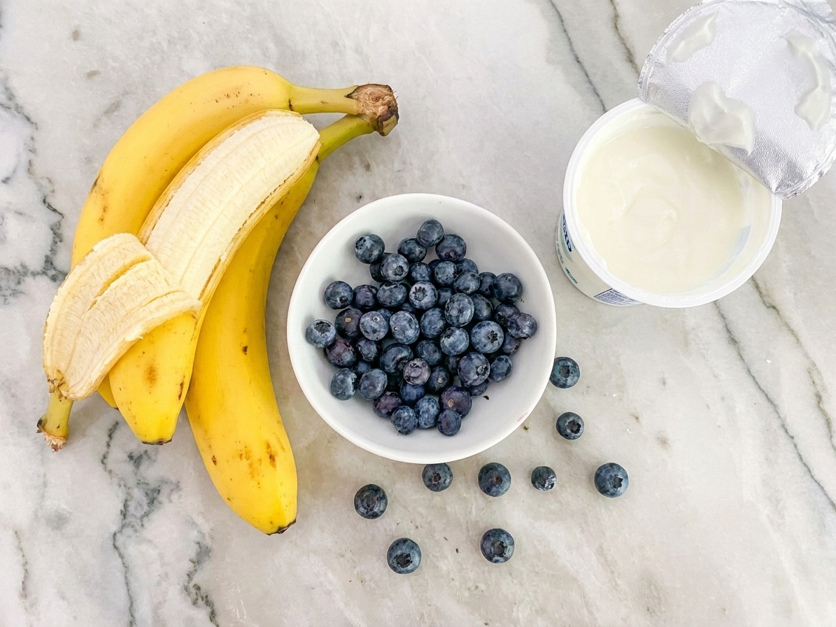 A small arrangement of ingredients on a light kitchen counter: ripe bananas, fresh blueberries in a bowl, and a container of plain unsweetened yogurt