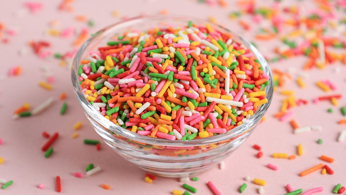 A small bowl filled with rainbow jimmies sprinkles on a baking counter next to measuring spoons