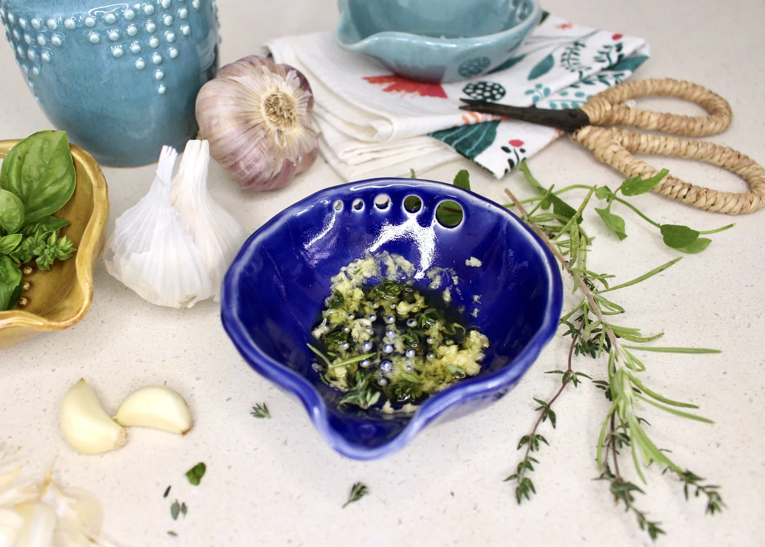 A small bowl of Dijon mustard, olive oil, minced garlic, chopped rosemary, thyme, salt, and pepper being mixed with a spoon on a kitchen counter