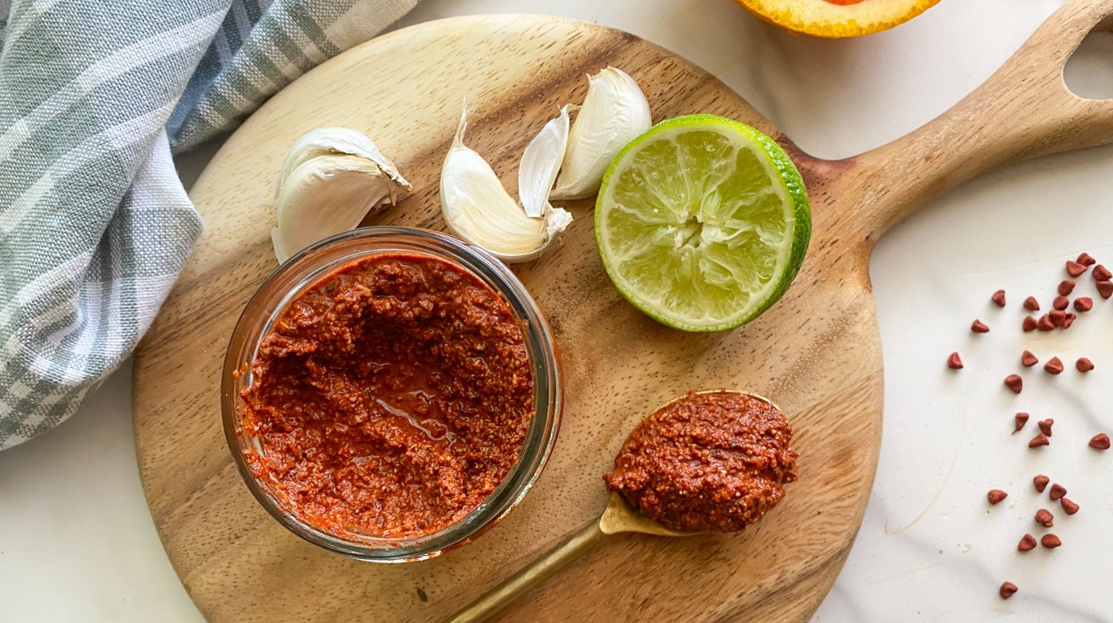 A small bowl of achiote paste with halved oranges and limes, peeled garlic cloves, and spices arranged on a light kitchen counter