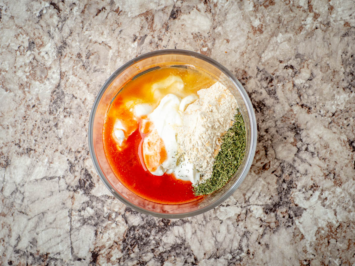 A small bowl of buffalo ranch dressing being whisked until smooth on a kitchen counter, with hot sauce and lemon nearby