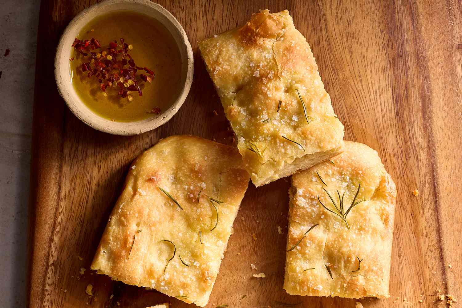 A small bowl of chopped rosemary, a dish of flaky salt, and a bottle of olive oil on a kitchen counter