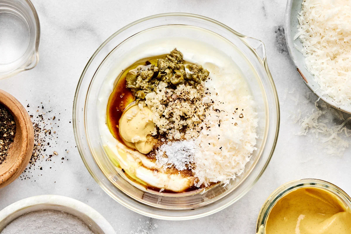 A small bowl of creamy Caesar dressing on a light stone counter with a spoon resting beside it, with a lemon wedge and Parmesan nearby