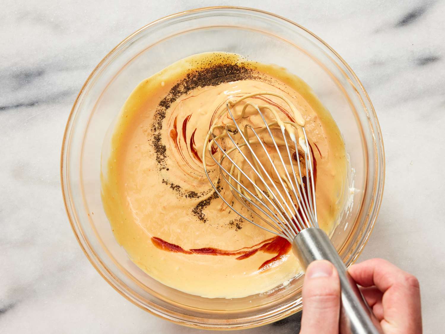 A small bowl of creamy burger sauce being stirred with a spoon on a kitchen counter, with pickles and condiments nearby in warm lighting