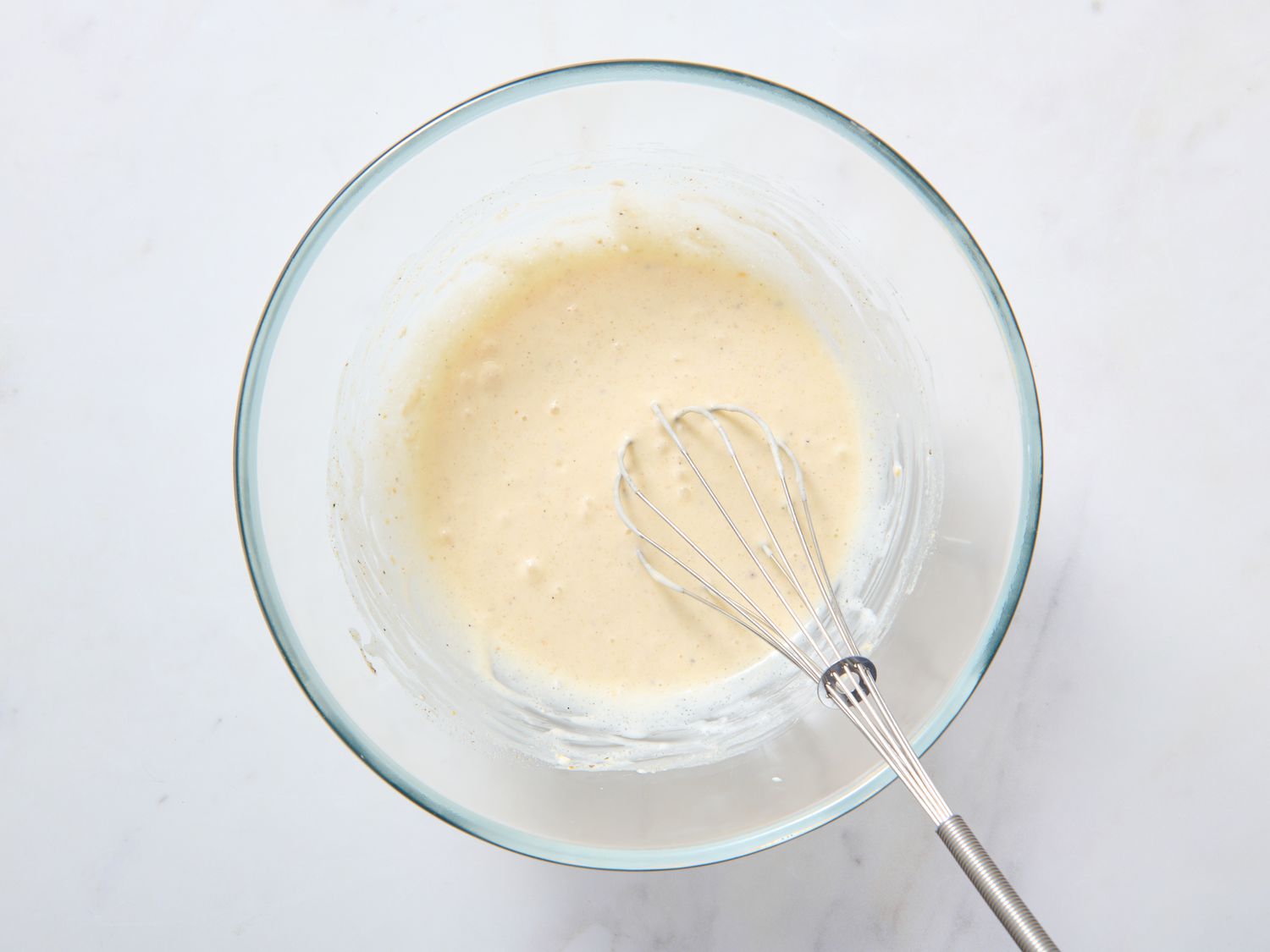 A small bowl of creamy coleslaw dressing on a light kitchen counter with a whisk and sliced cabbage nearby, natural window light