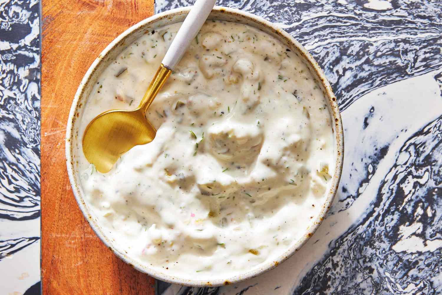 A small bowl of creamy homemade tartar sauce with visible pickle and herb flecks on a wooden table next to lemon wedges and a plate of crispy fish fillets