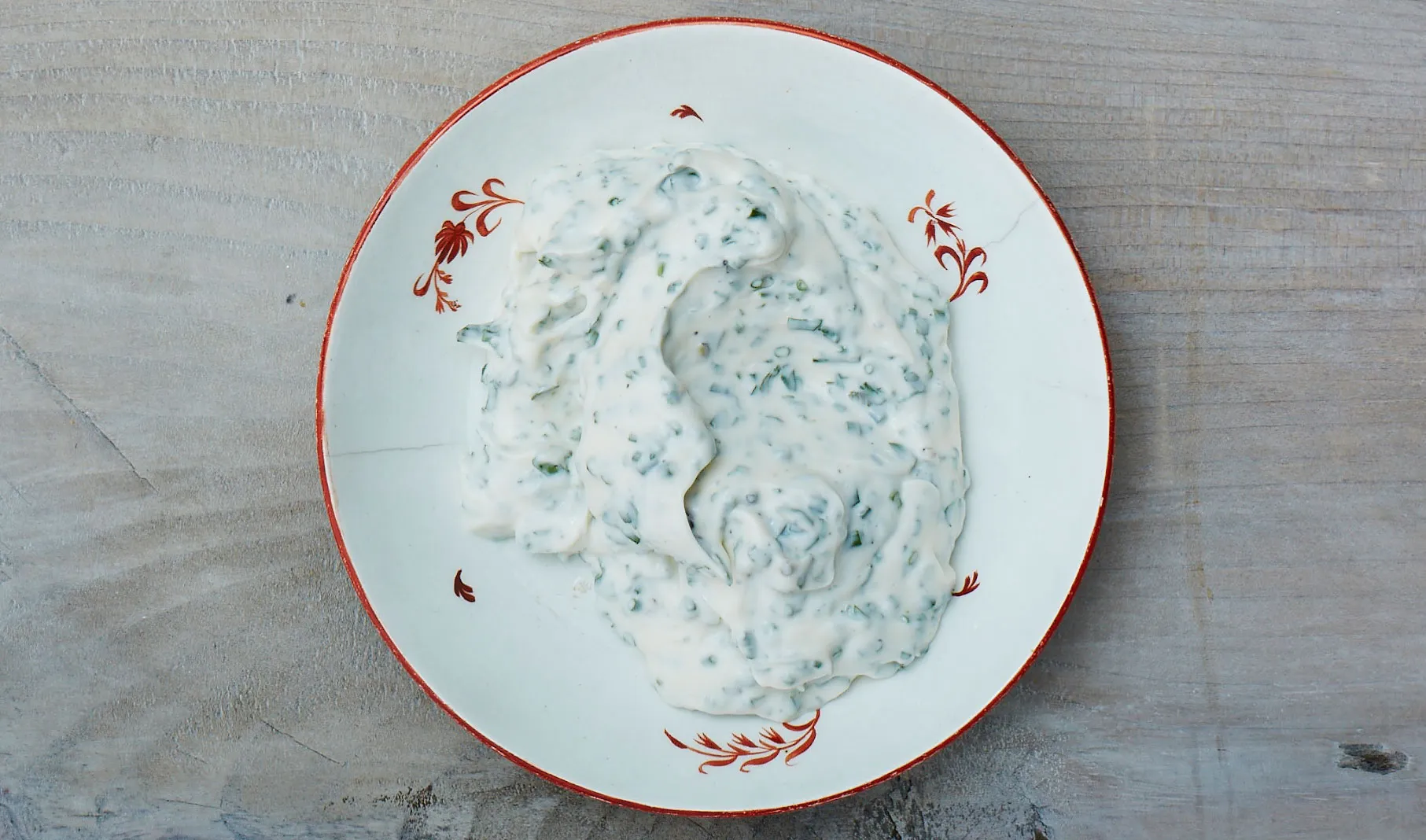 A small bowl of creamy lemon herb mayonnaise with visible dill and parsley on a kitchen counter beside a lemon and a microplane