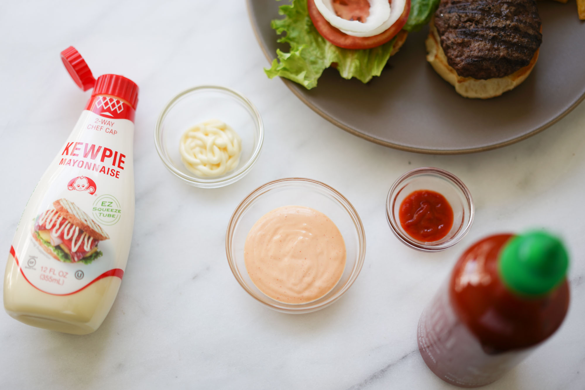 A small bowl of creamy sriracha mayo being whisked with a spoon on a kitchen counter next to lime and soy sauce