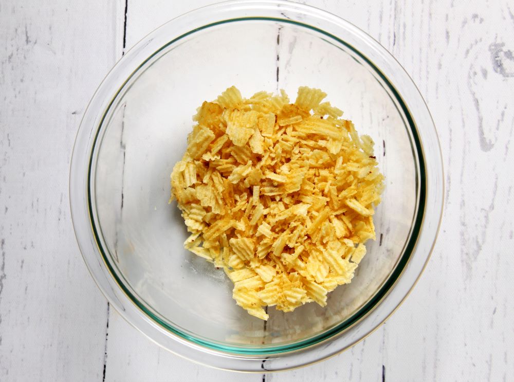 A small bowl of crushed plain potato chips on a kitchen counter next to chopped chives