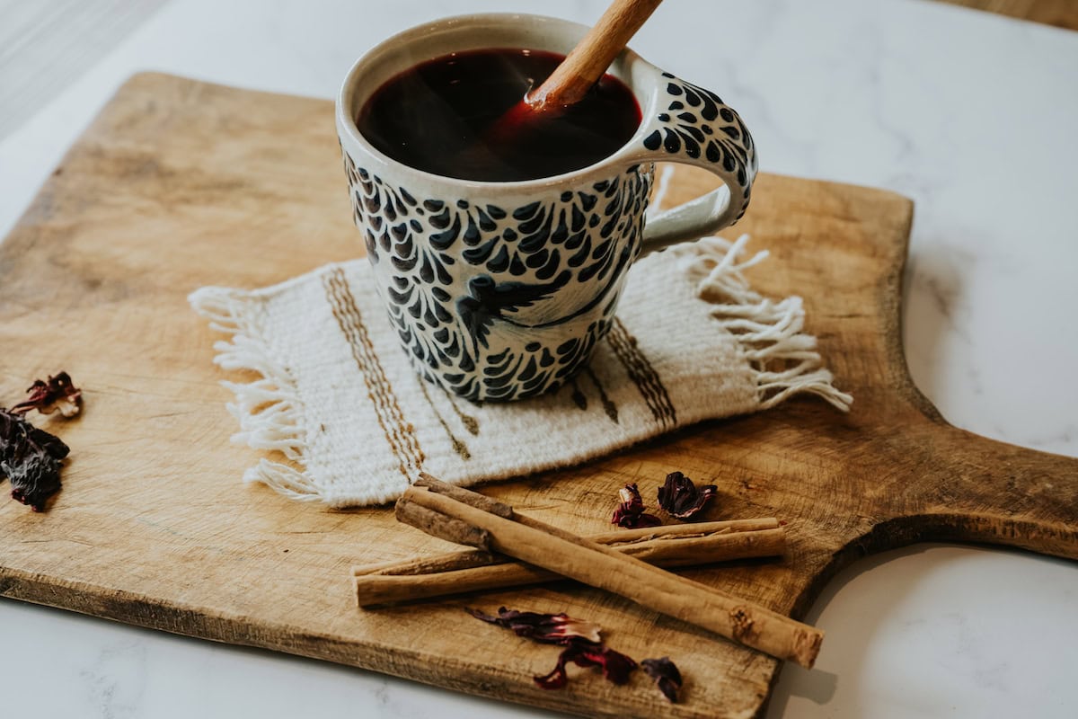 A small bowl of dried hibiscus flowers next to a cinnamon stick, limes, and a measuring cup on a clean kitchen counter