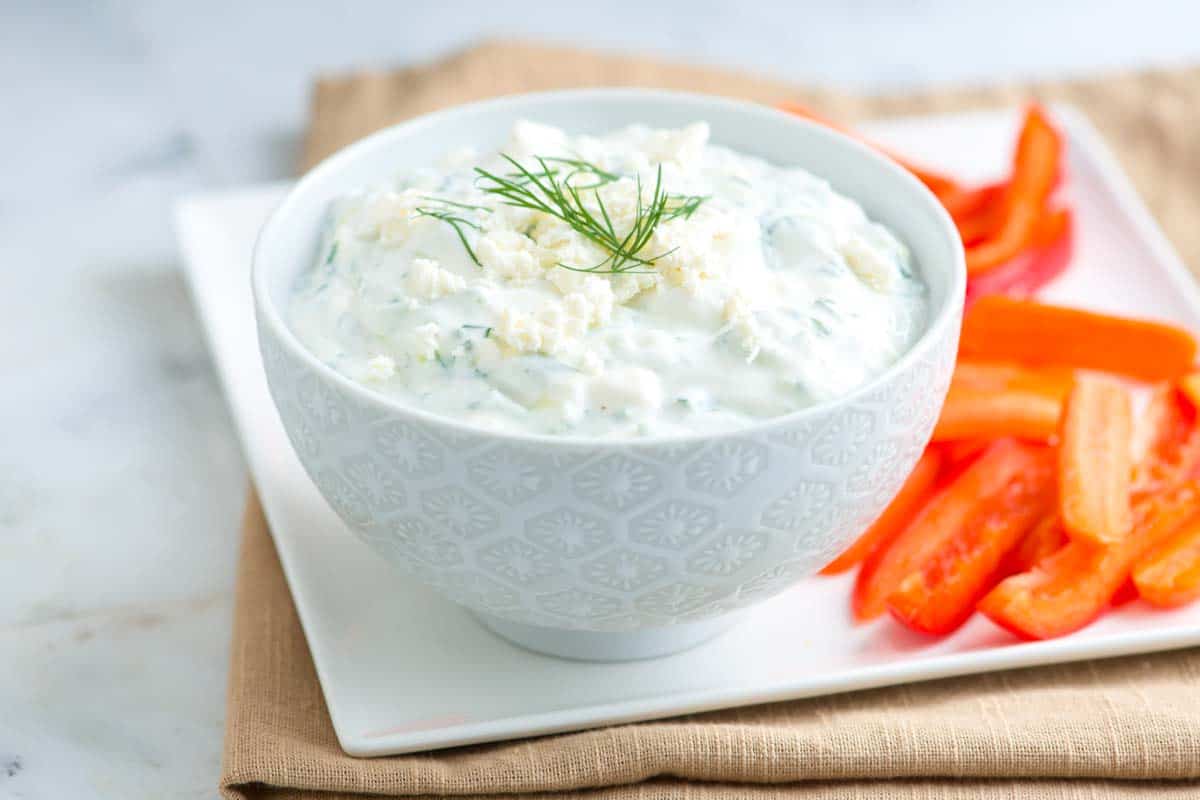 A small bowl of feta yogurt sauce being stirred with a spoon on a wooden cutting board next to a lemon and fresh herbs