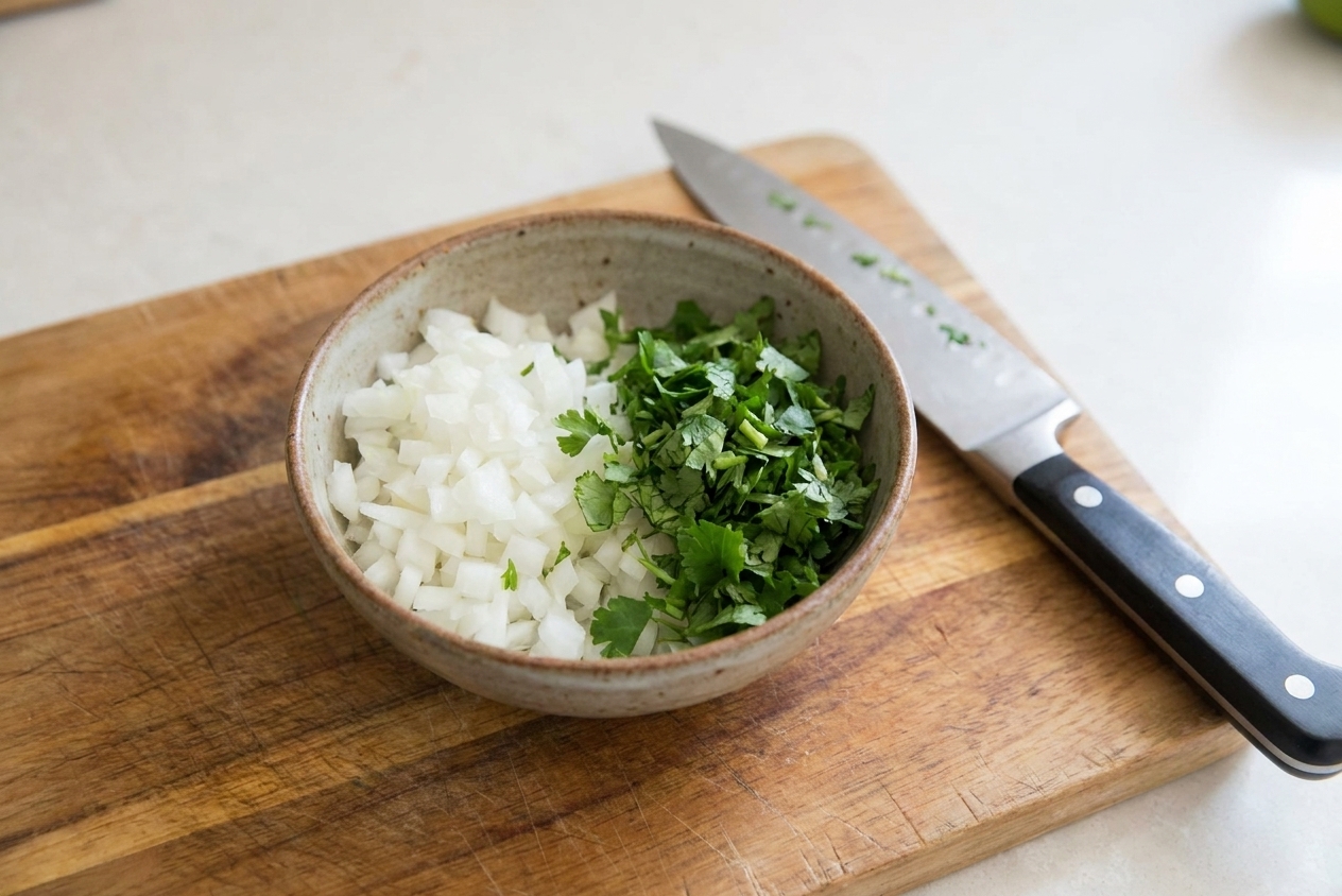 A small bowl of finely diced white onion and chopped cilantro on a cutting board with a chef's knife nearby