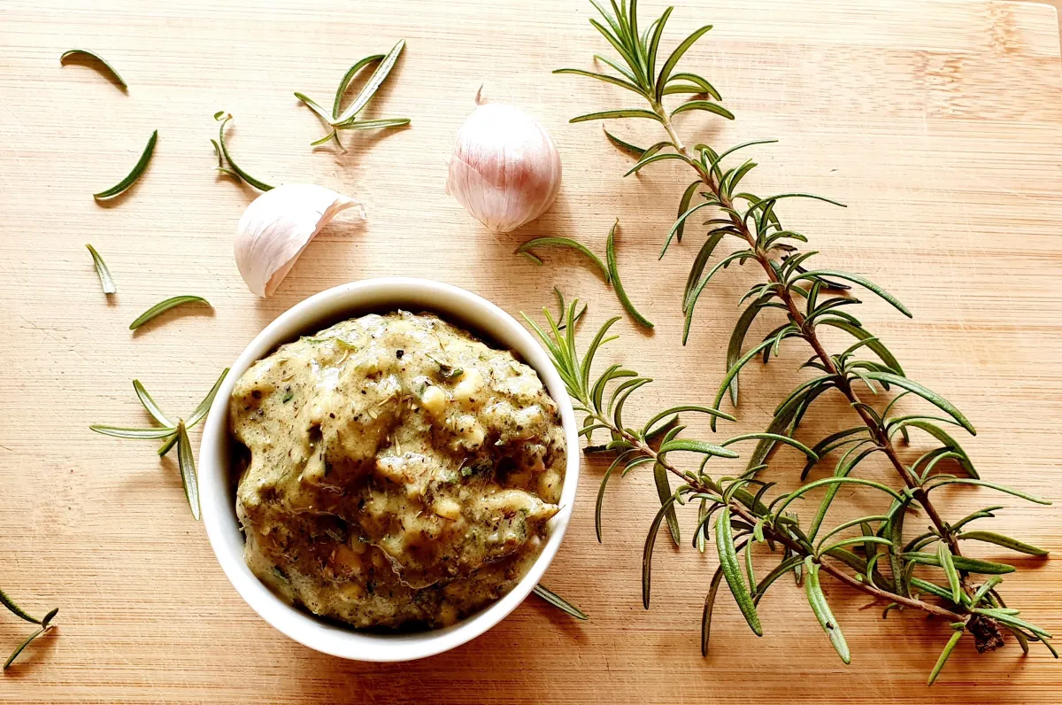A small bowl of garlic herb Dijon rub with a spoon beside it on a kitchen counter, with rosemary and thyme scattered nearby