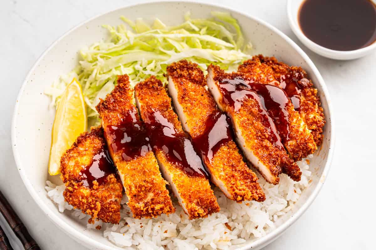 A small bowl of glossy brown tonkatsu sauce on a light kitchen counter with a spoon resting beside it, warm natural light