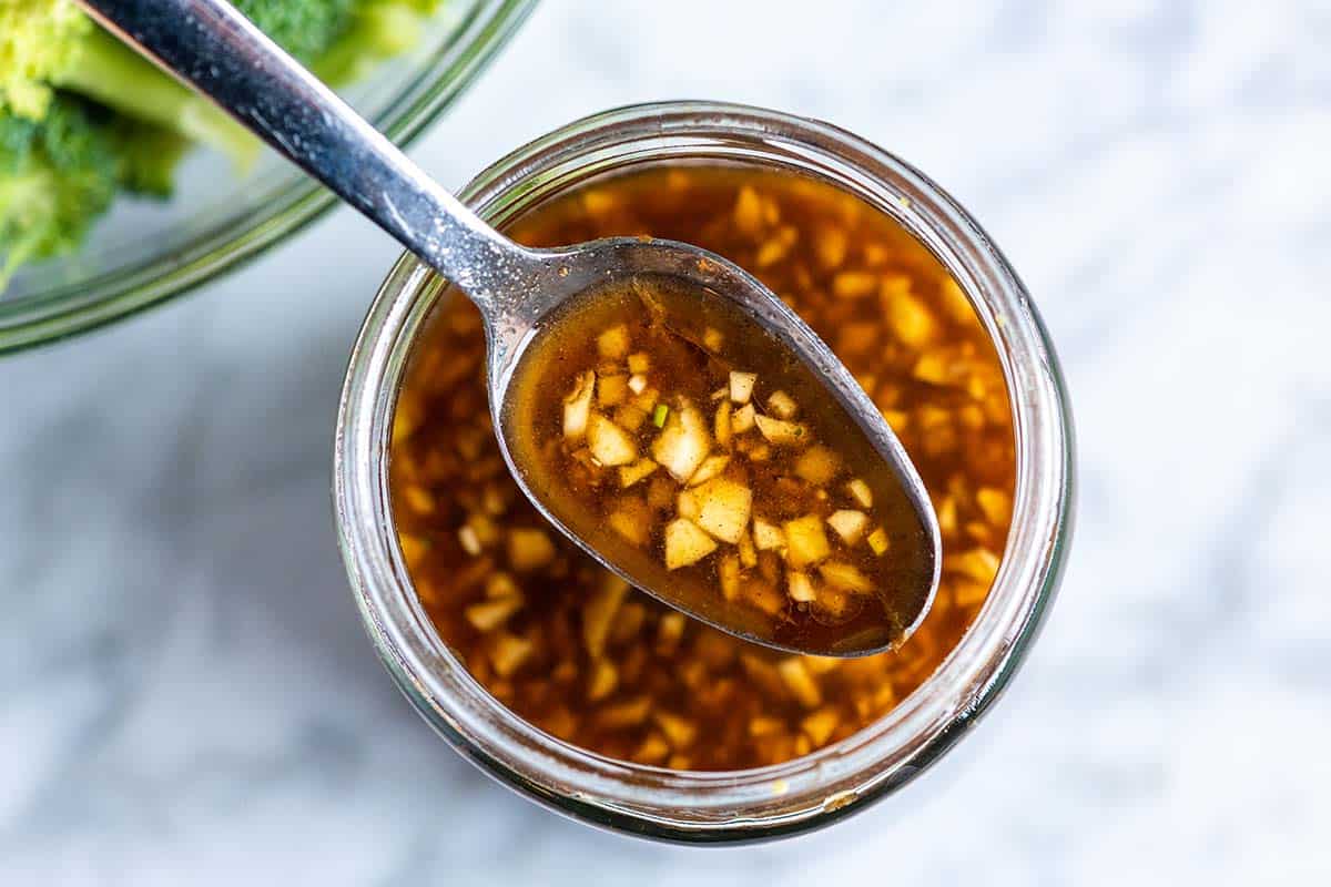 A small bowl of glossy ginger-garlic sauce with soy sauce, lime, and sriracha on a kitchen counter next to a whisk