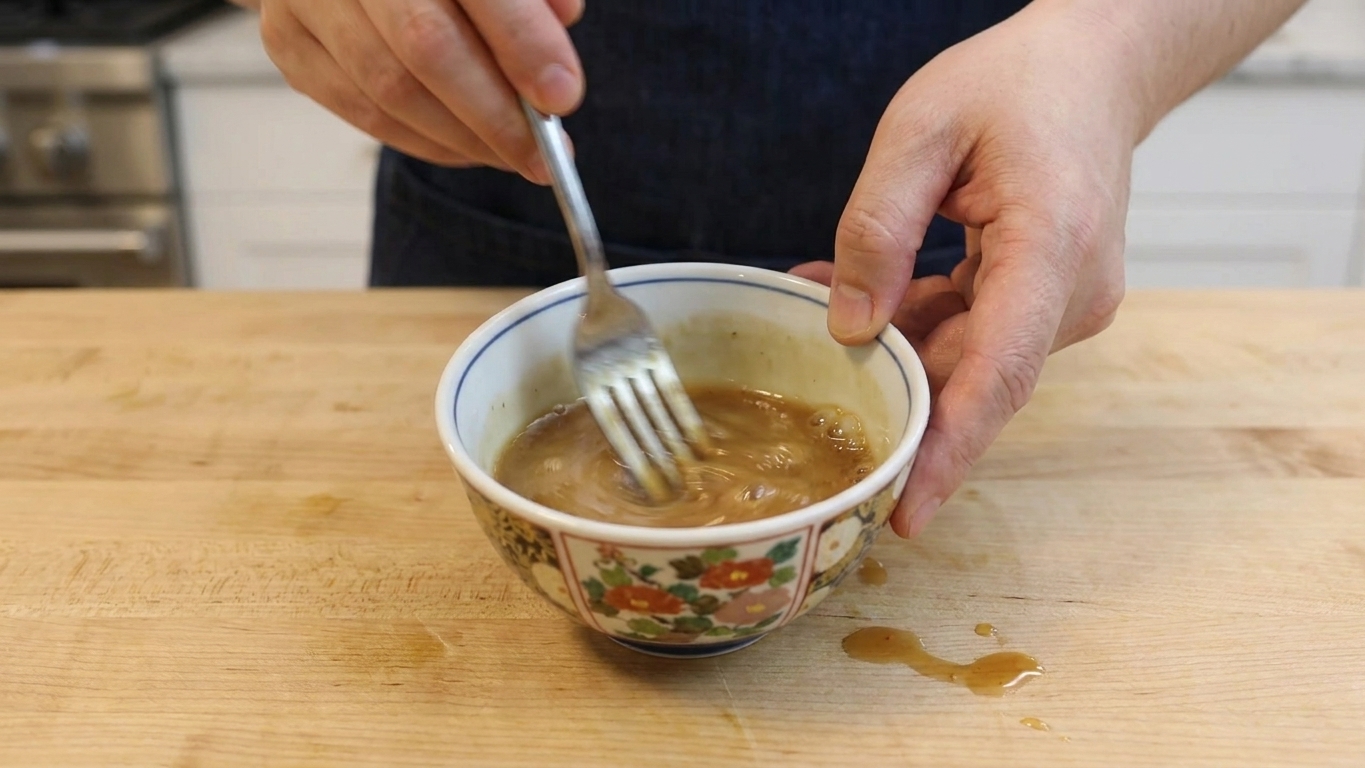 A small bowl of light brown stir-fry sauce being whisked with a fork on a wooden counter