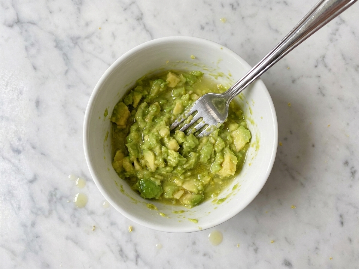 A small bowl of mashed avocado with visible chunks and lemon juice being mixed with a fork on a kitchen counter