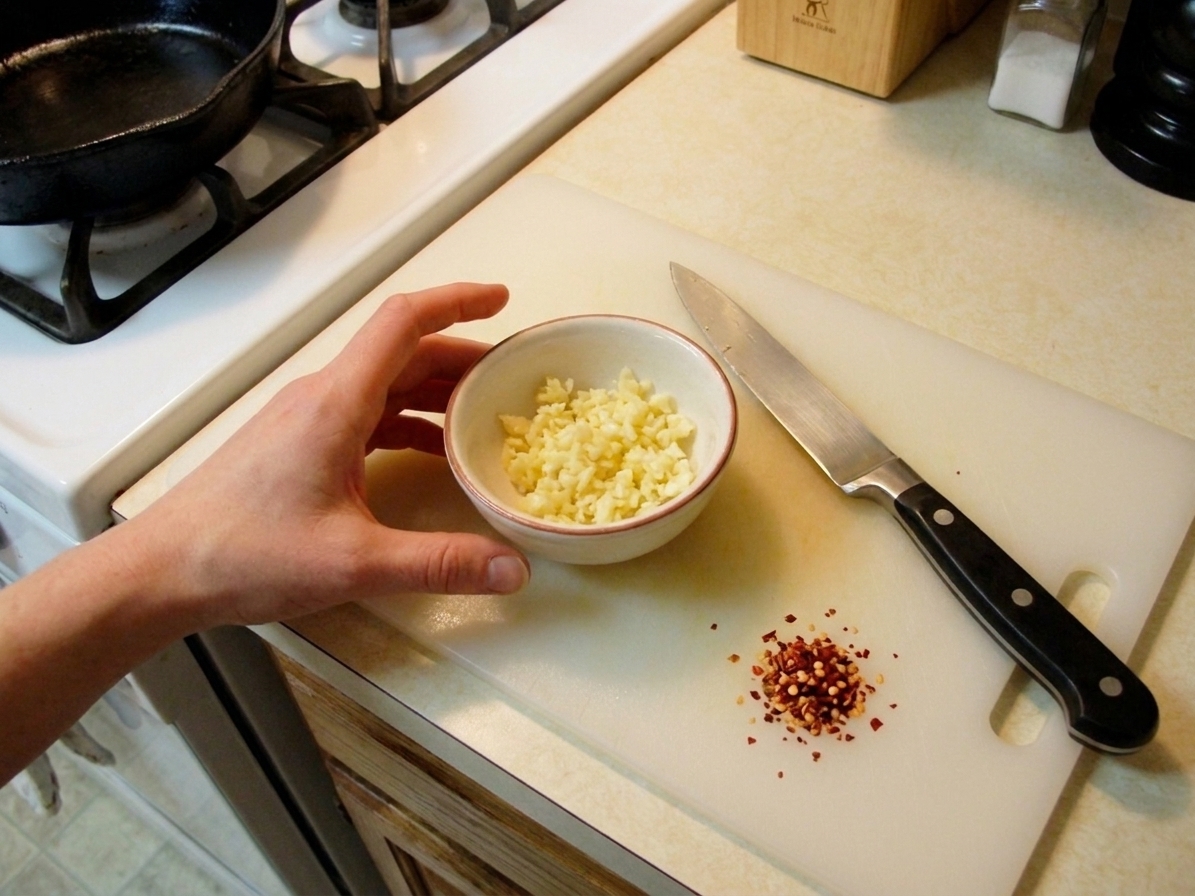 A small bowl of minced garlic and red pepper flakes on a kitchen counter next to a chef's knife