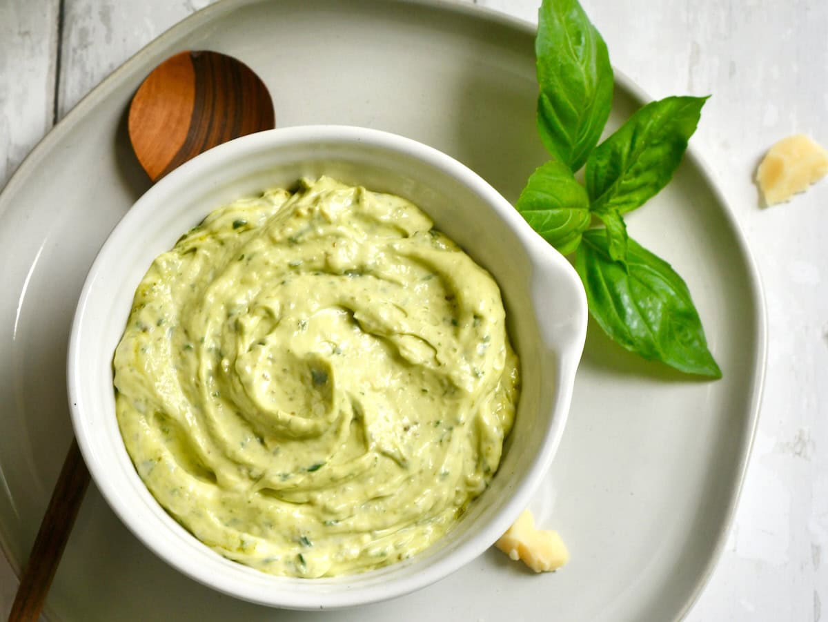 A small bowl of pale green pesto mayonnaise on a wooden kitchen counter with a spoon beside it, with a burger bun and sliced tomato softly blurred in the background