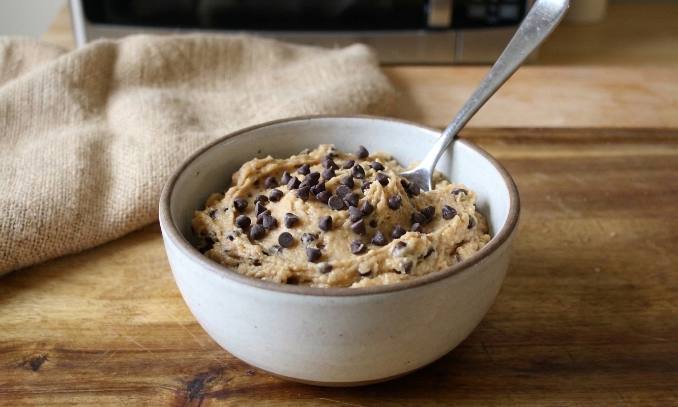 A small bowl of protein cookie dough topped with mini chocolate chips on a simple table setting with a spoon