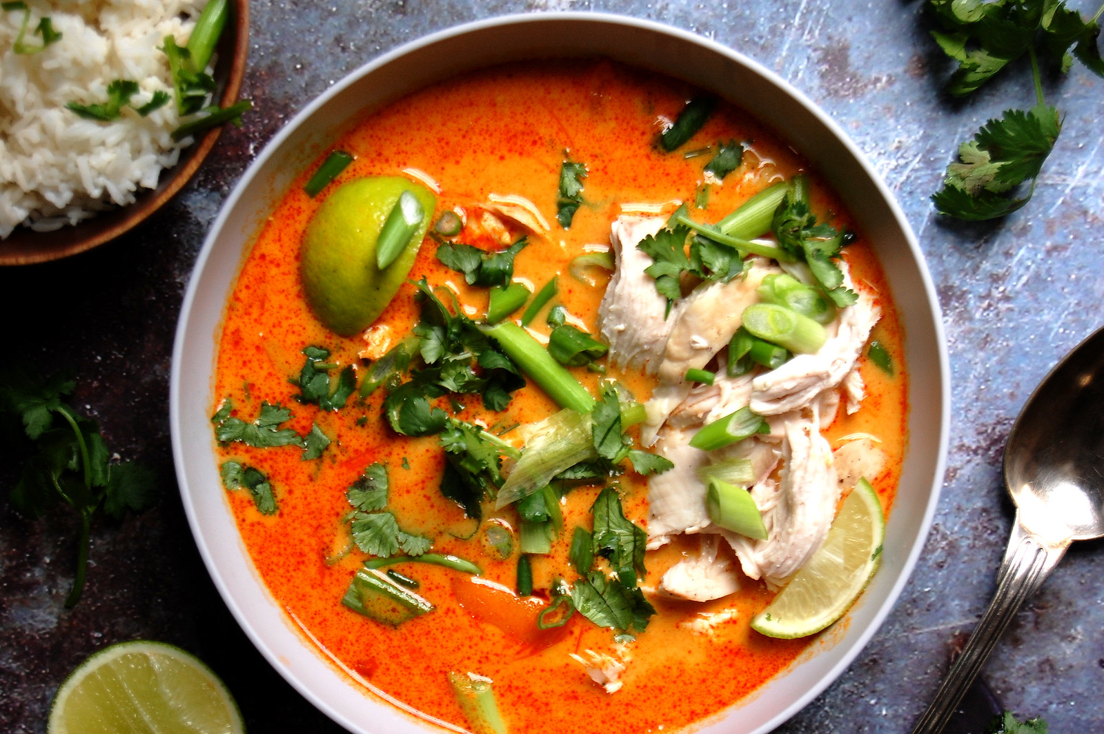 A small bowl of red curry paste next to an opened can of coconut milk and a cut lime on a kitchen counter