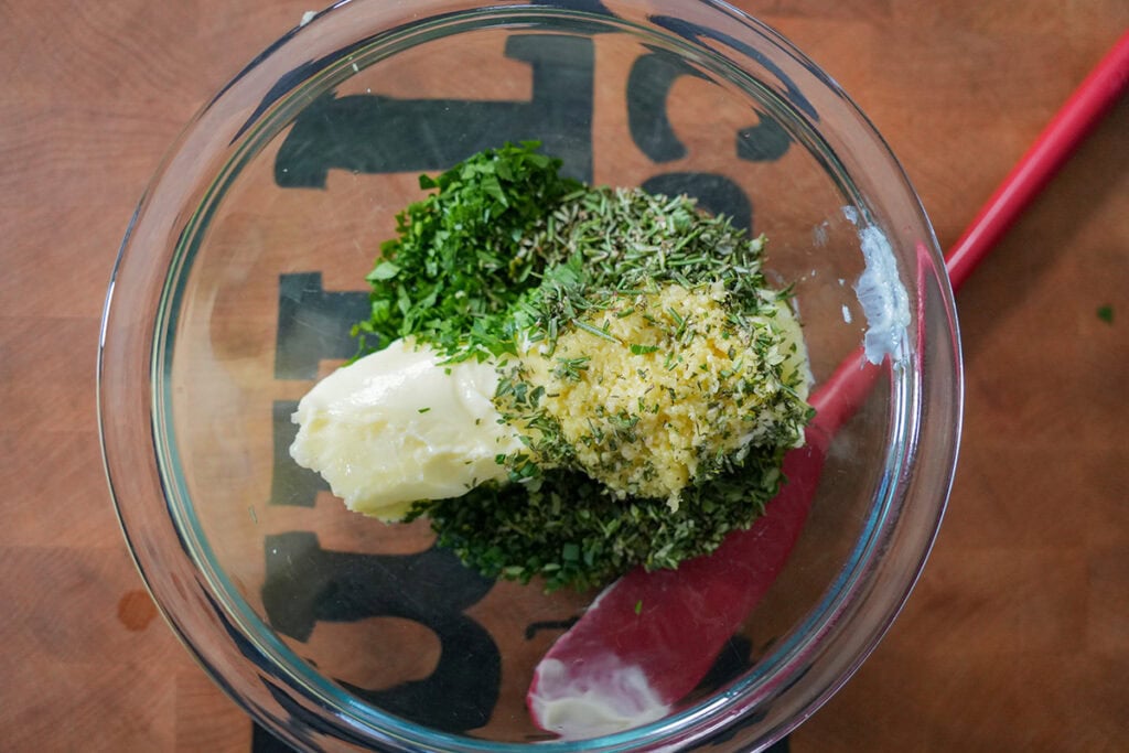 A small bowl of softened butter mixed with chopped herbs, garlic, and lemon zest on a wooden cutting board with a spoon nearby