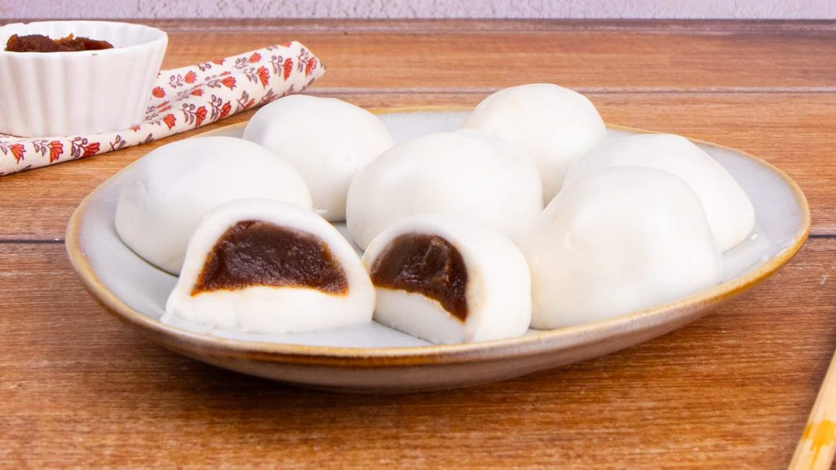A small bowl of sweet red bean paste with a spoon on a kitchen counter next to a tray dusted with starch