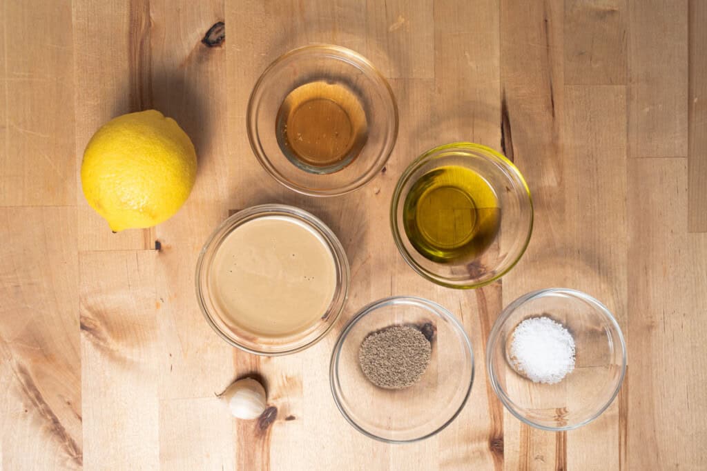 A small bowl of tahini with a lemon, garlic clove, honey, salt, and a measuring spoon of warm water on a kitchen counter