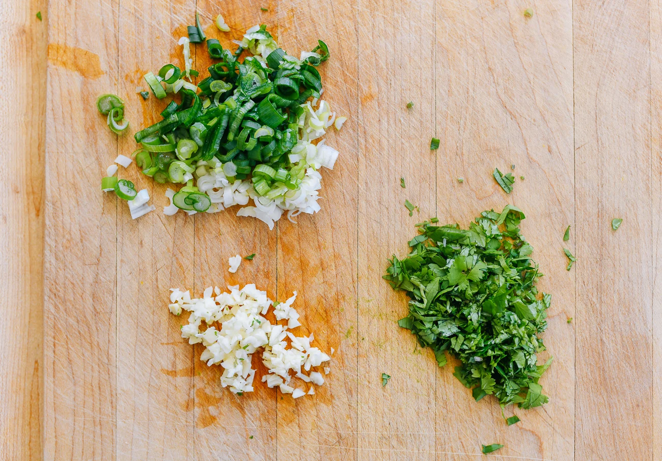 A small bowl with minced garlic and thinly sliced scallions on a cutting board beside a chef knife