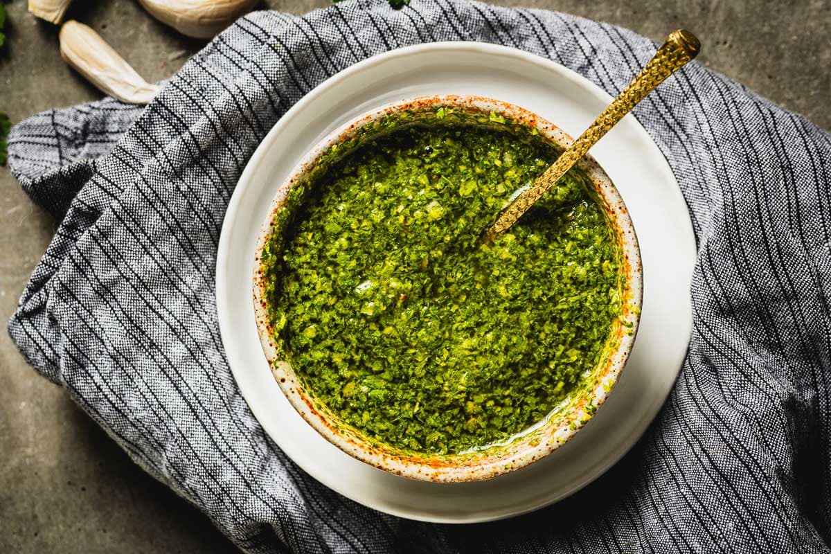 A small ceramic bowl filled with bright green chimichurri sauce, with a spoon resting beside it on a clean kitchen counter under warm natural light