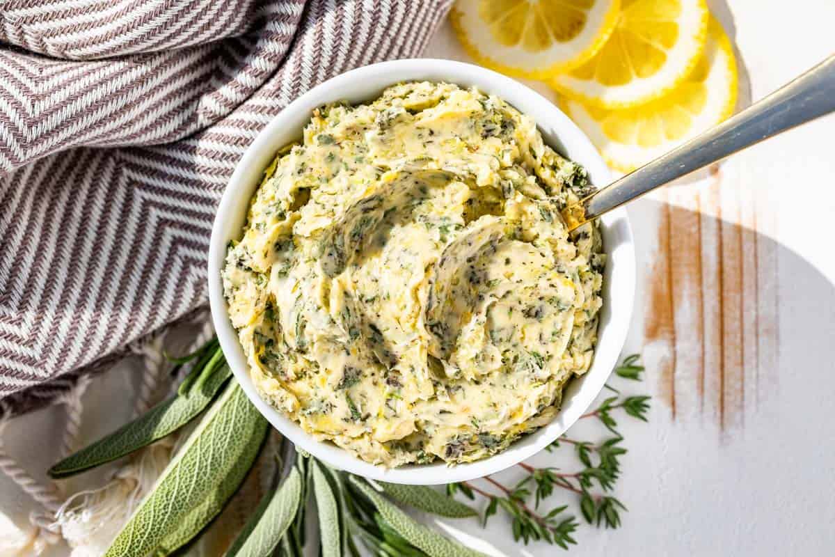 A small ceramic bowl filled with garlic herb butter flecked with chopped rosemary, thyme, and parsley on a kitchen counter next to a microplane and lemon