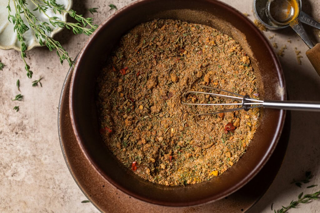 A small ceramic bowl filled with homemade jerk seasoning on a kitchen counter, surrounded by measured spices and a wooden spoon