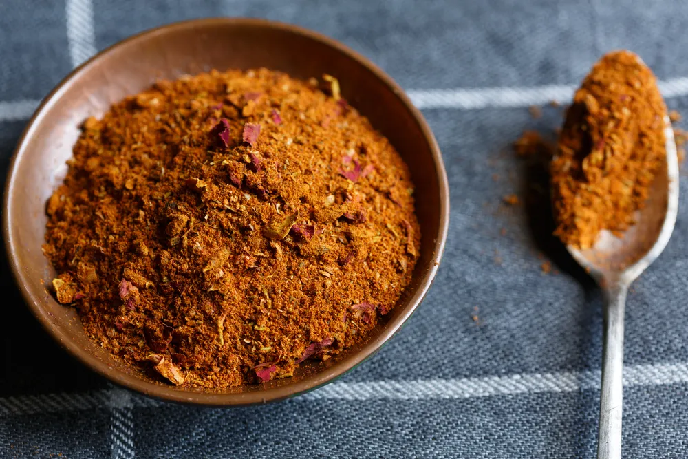 A small ceramic bowl filled with ras el hanout spice blend beside measuring spoons on a light stone kitchen counter
