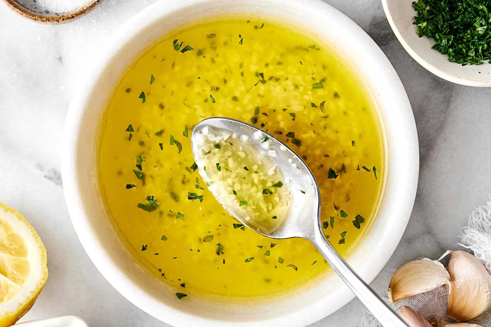 A small ceramic bowl with melted butter, olive oil, minced garlic, and lemon zest being stirred with a spoon on a light kitchen counter