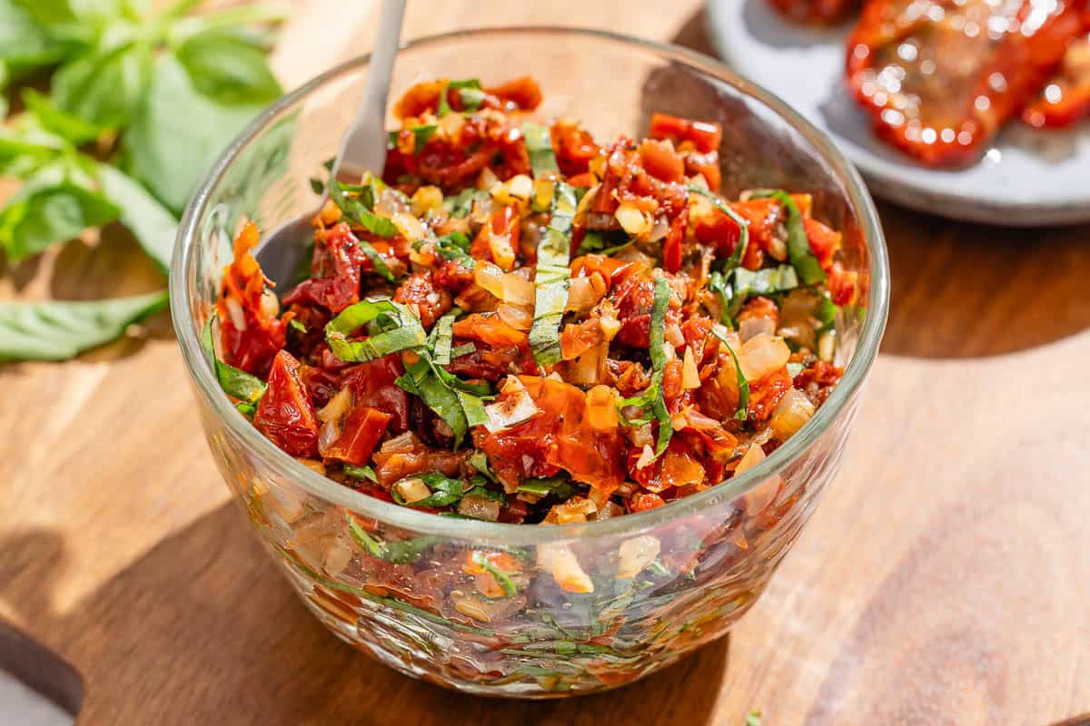 A small cutting board with sliced sun-dried tomatoes, minced garlic, and chopped onion next to a jar of sun-dried tomatoes on a kitchen counter