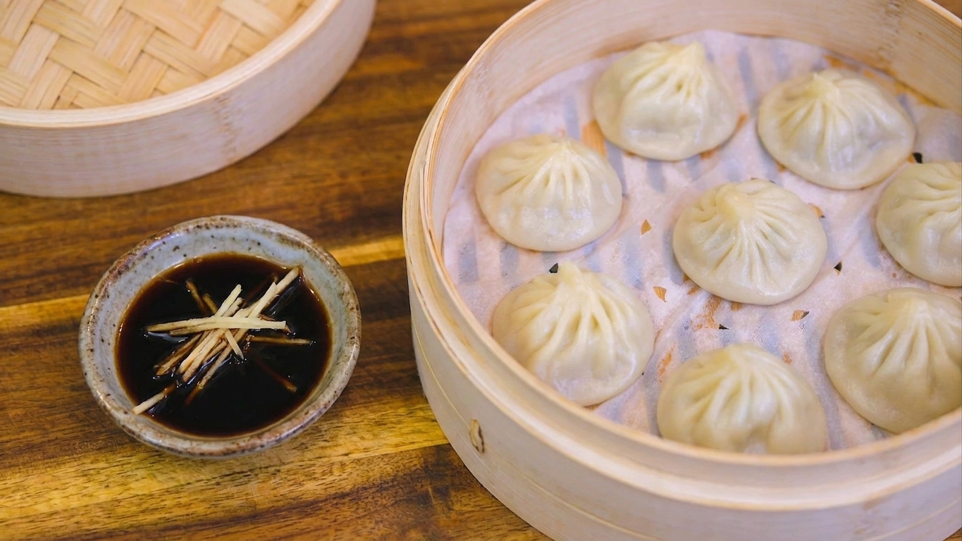 A small dipping dish of black vinegar topped with thin ginger matchsticks beside a plate of steamed xiao long bao.