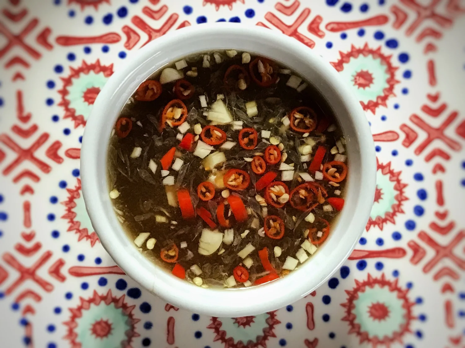 A small glass bowl of Vietnamese nuoc cham with visible minced garlic and sliced red chile on a wooden table next to lime wedges
