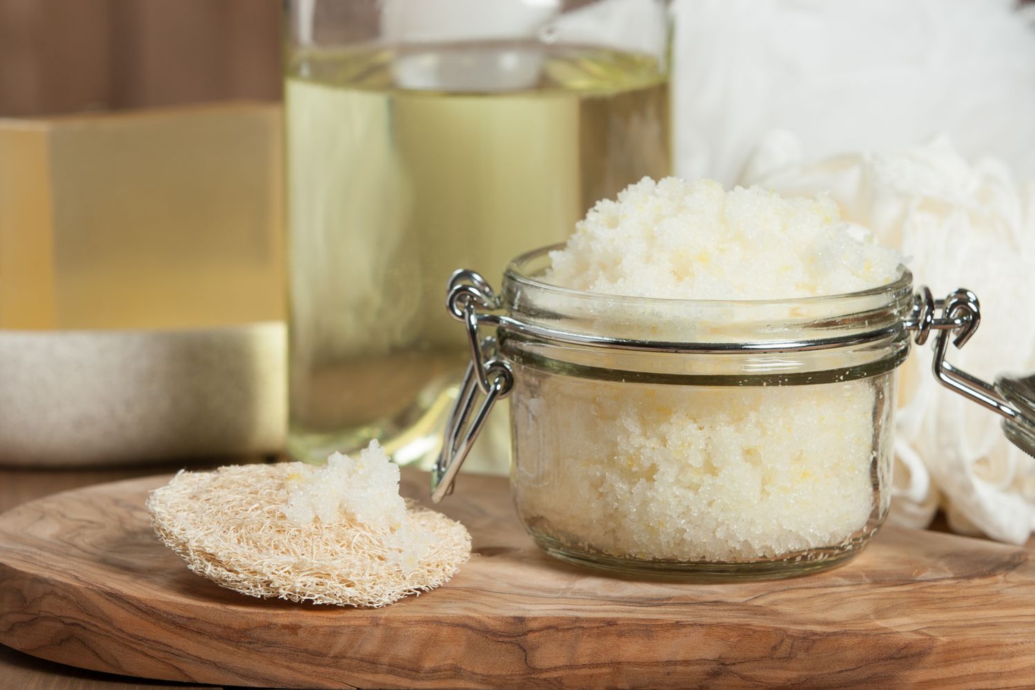 A small glass jar filled with brown sugar scrub on a warm-toned kitchen counter, with a wooden spoon and a folded towel nearby, natural window light