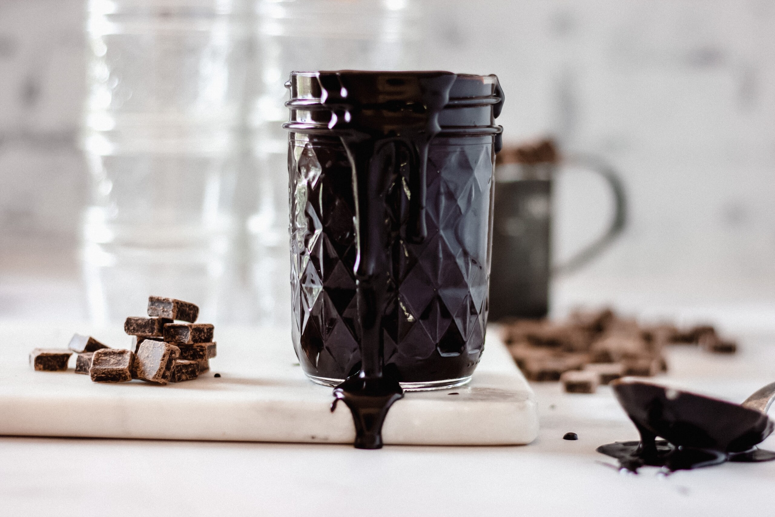 A small glass jar filled with cooled hot fudge sauce sitting on a kitchen counter next to a spoon