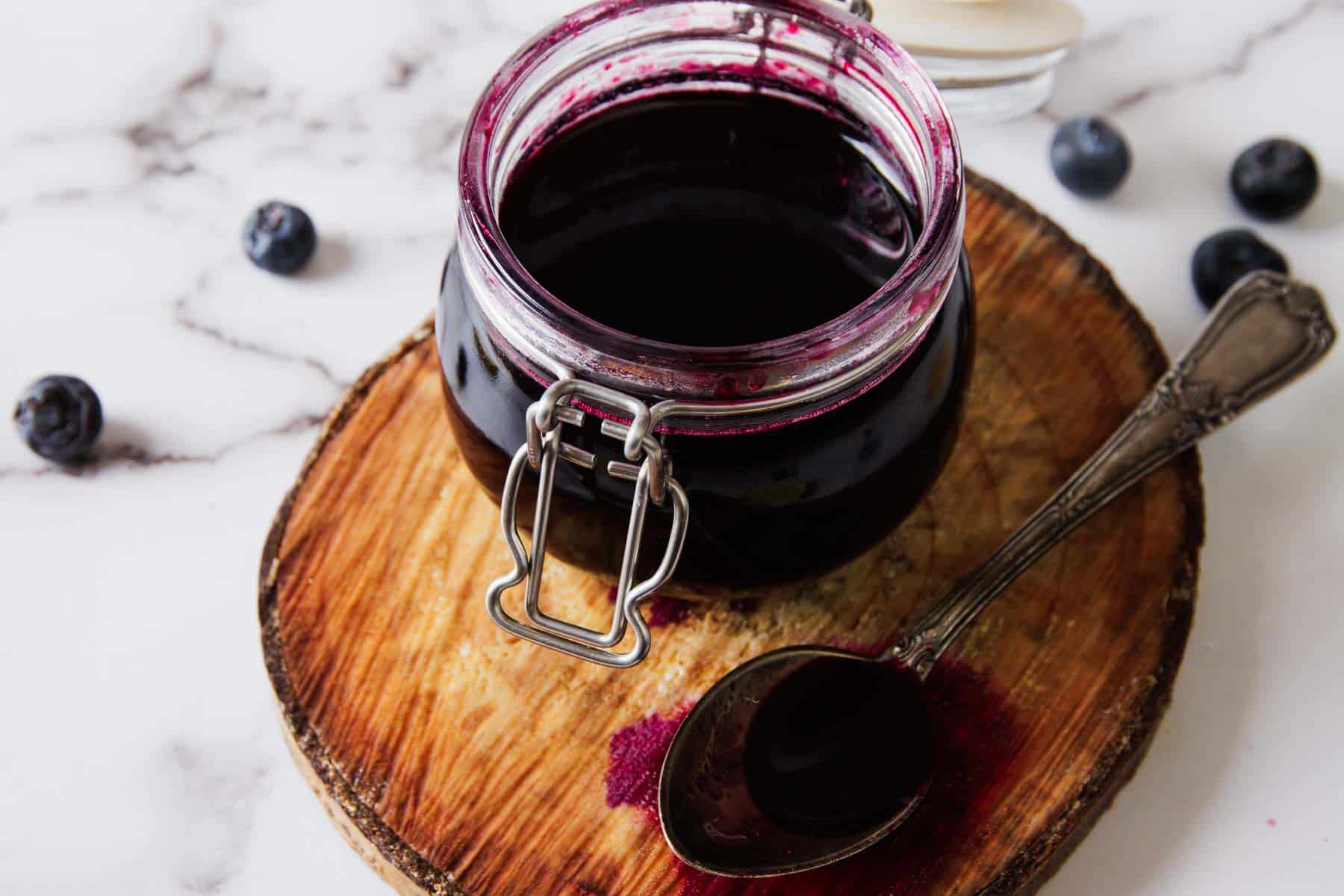 A small glass jar filled with deep purple blueberry syrup sitting on a kitchen counter with a spoon beside it