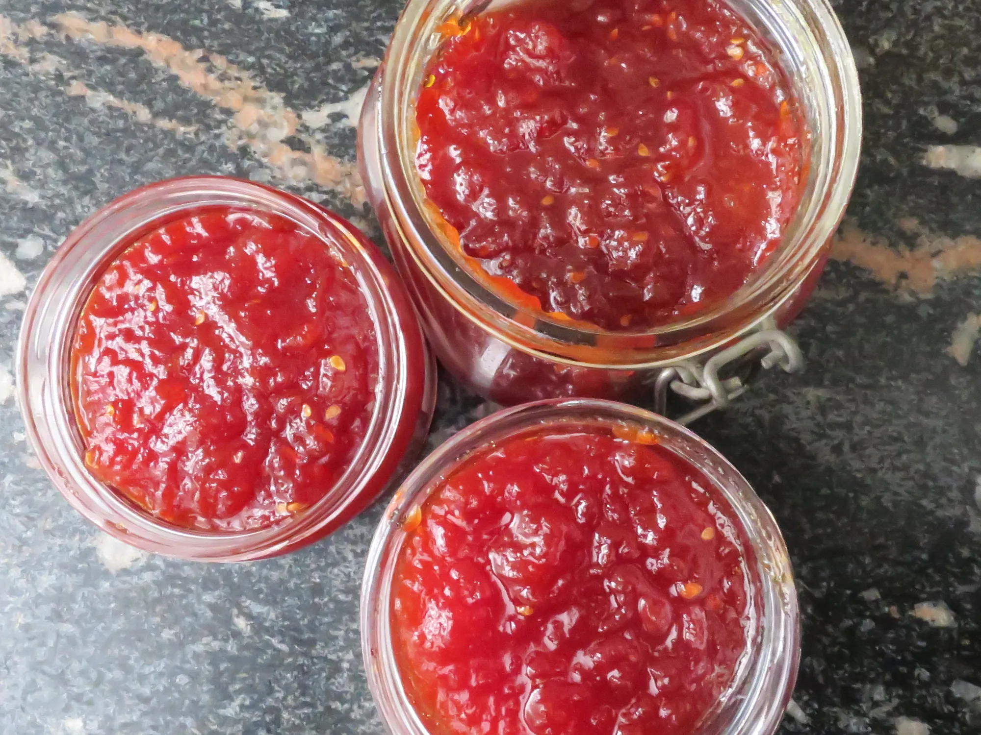 A small glass jar filled with glossy deep red tomato jam on a light kitchen counter, with a spoon resting beside it and soft natural window light