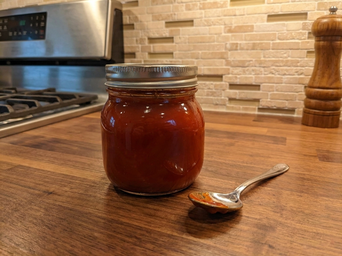 A small glass jar filled with glossy homemade ketchup on a warm kitchen counter with a spoon beside it