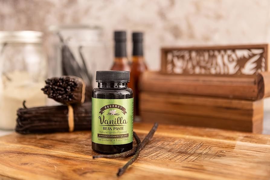 A small glass jar filled with glossy homemade vanilla bean paste with visible vanilla specks on a light kitchen counter, a spoon resting beside it
